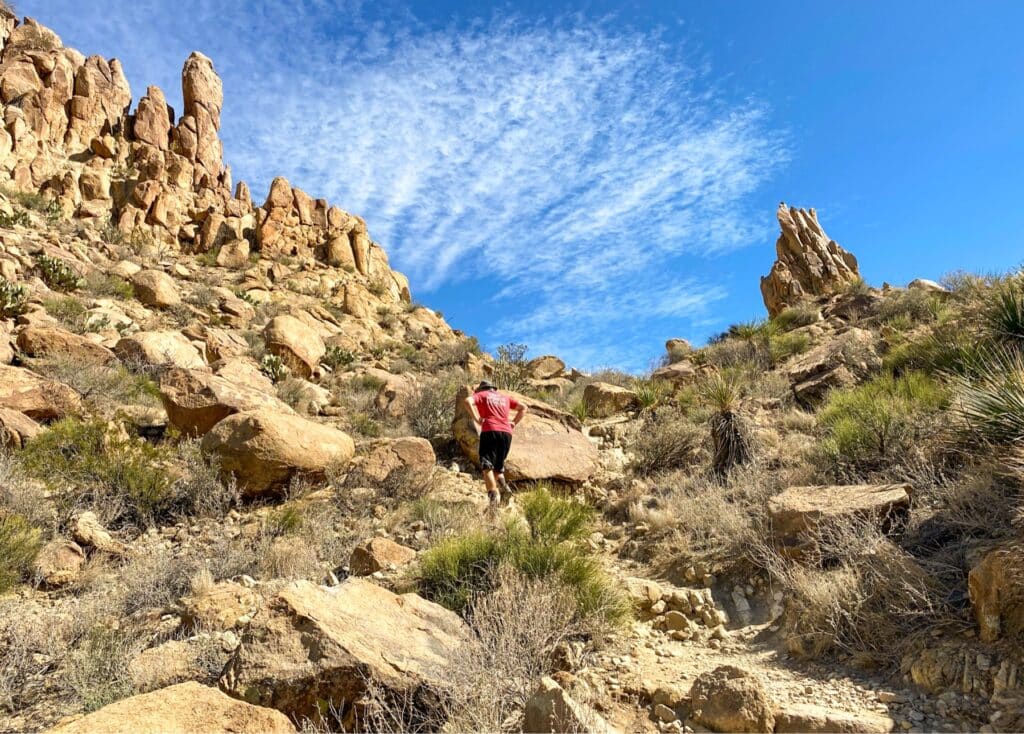 A man is climbing the boulders on Grapevine Hills trail in big bend National park. He's wearing a red shirt and black shorts and is looking at the ground with both hands on his hips.