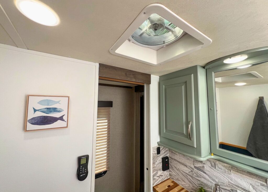 Inside an RV bathroom, a white Maxxfan is open and running. On the left, a black Maxxfan remote is mounted to the wall.
