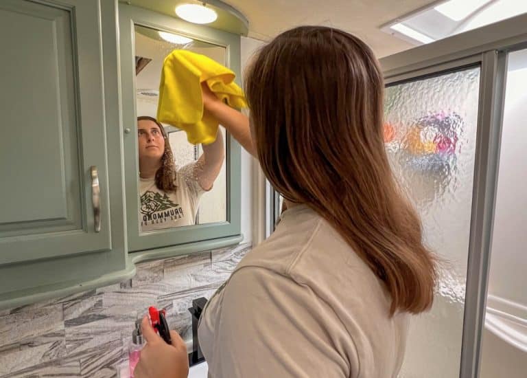 A woman is in an RV bathroom cleaning a mirror with a yellow microfiber cloth.