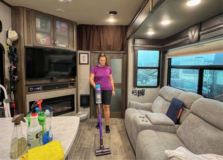 A woman wearing a purple shirt and black shorts is vacuuming inside of a fifth wheel RV using a Dyson stick vacuum. To the right a grey sofa sits in a slide-out. A TV is on the left and a variety of cleaning supplies sit on the kitchen counter.