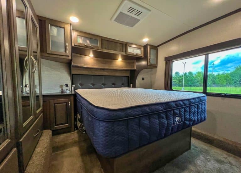Fifth wheel RV bedroom. Brown cabinets surround the bed on the left and above. A window is to the left of the bed. The mattress is in the middle and is bare with no bedding on it.
