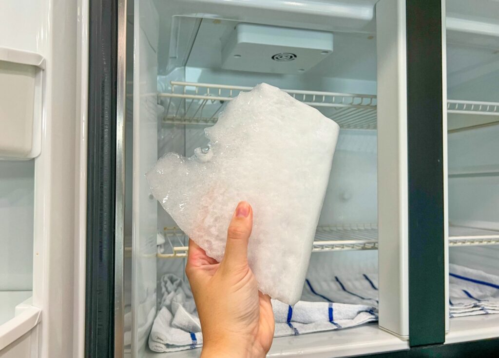 A hand is holding a large chunk of ice in front of an open RV freezer. Inside the freezer the back wall is filled with ice and a towel lays at the bottom of the freezer.