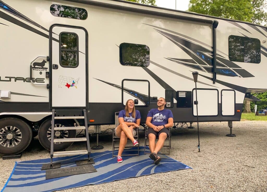 A man and a woman sit in chairs outside of their Dutchmen Voltage fifth wheel. They're relaxed, laughing, and enjoying the campsite.