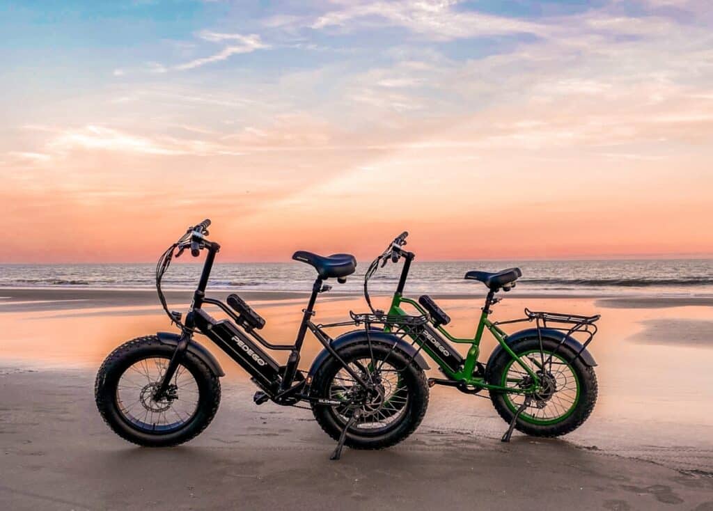 Two Pedego e-bikes parked on the beach at sunset. One bike is black, the other is green.