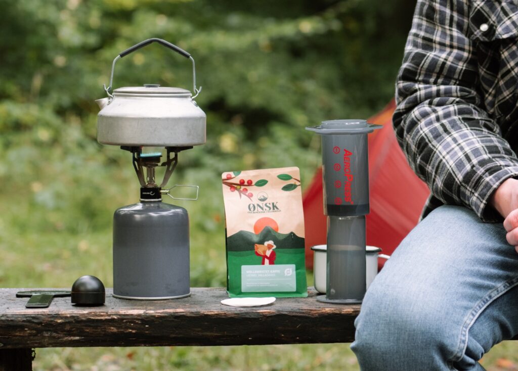 A person sits on a wood bench in front of their camping tent along with their coffee supplies. On the bench is kettle, bag of coffee, scoop, mug, and AeroPress.