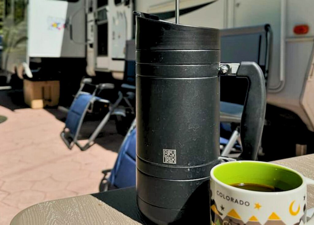 A black BruTrek French press sits on a table next to a Starbucks Coffee mug that says "Colorado". In the background is a white Montana fifth wheel and two blue zero gravity camp chairs.