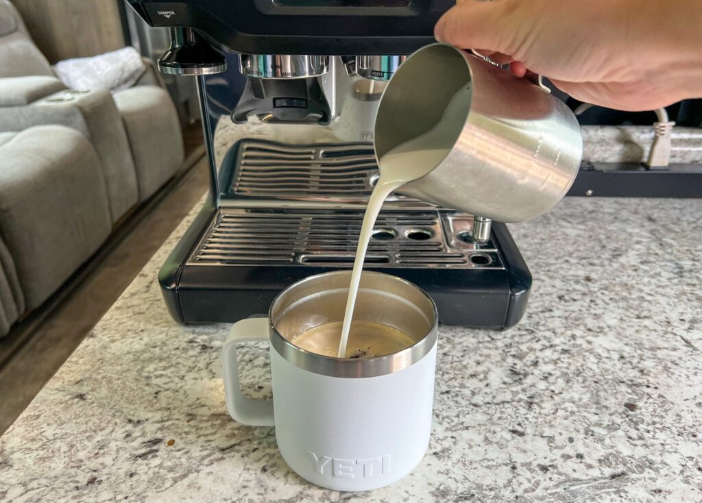 Hand is pouring frothed milk from a stainless steel pitcher and into a white yeti mug filled with coffee. In the background, an espresso machine sits on the counter top in an RV.