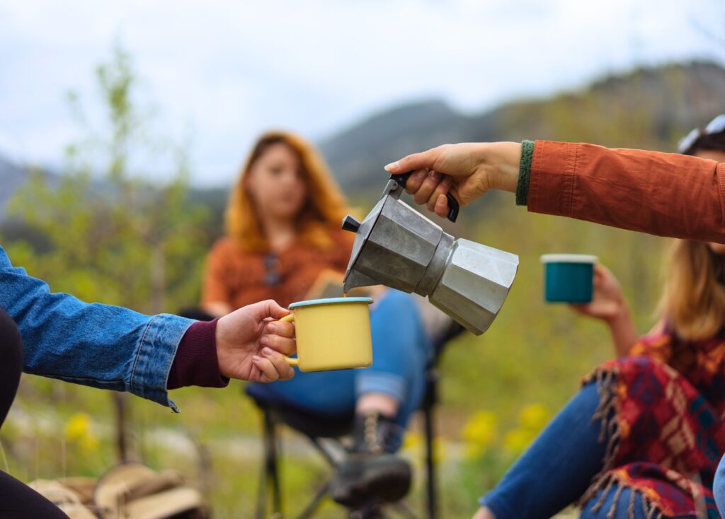 Group of friends sitting in camp chairs. A hand is reaching out to pour coffee from a moka pot into a mug another person is holding out.