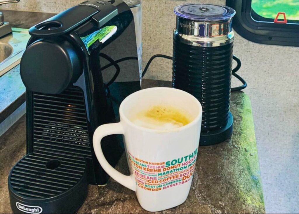 DeLonghi Nespresso single serve coffee maker sitting on counter in RV. A white coffee mug and milk frother sit next to it.