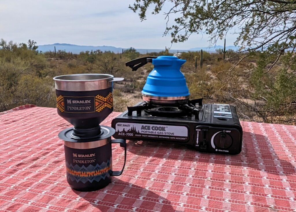 A Pendleton version of the Stanley Pour Over sits on a table covered with a red picnic table cover. Behind the pour over coffee mug is a portable camp stove with a blue collapsible kettle sitting on top of it.