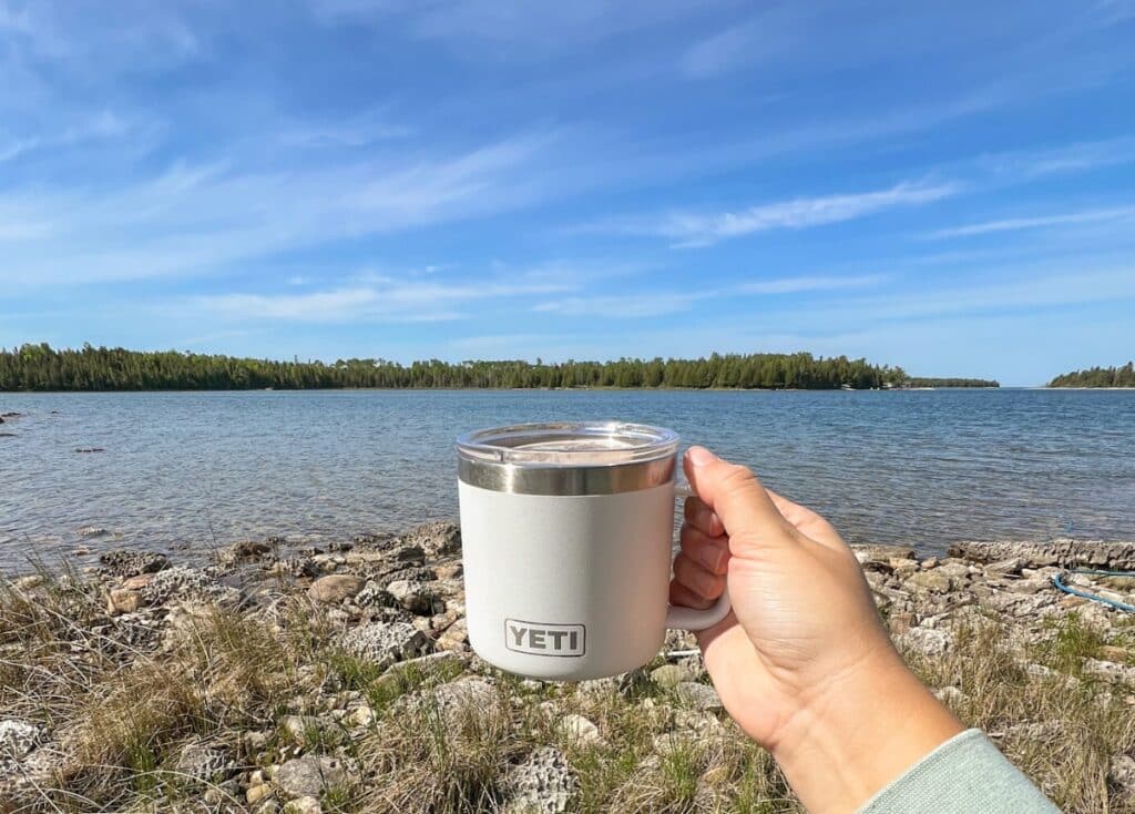 A hand holds up a white Yeti coffee mug in front of a lake. 