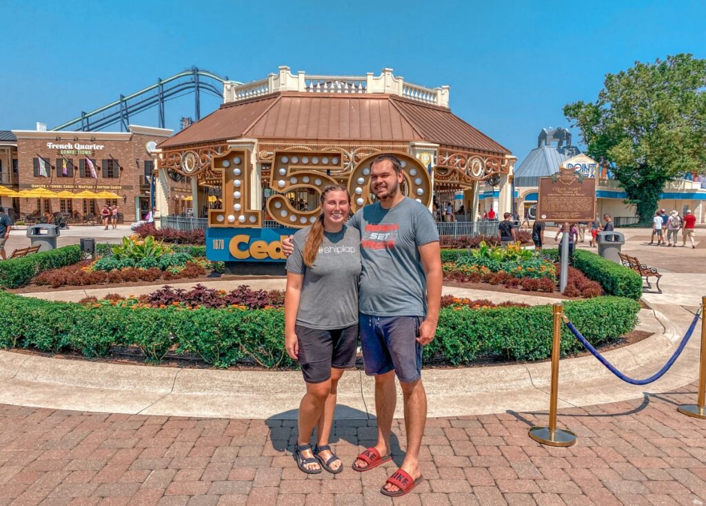 Megan and Philip standing in front of a 150 year anniversary Cedar Point Sign in front of the carousel at the amusement park. 