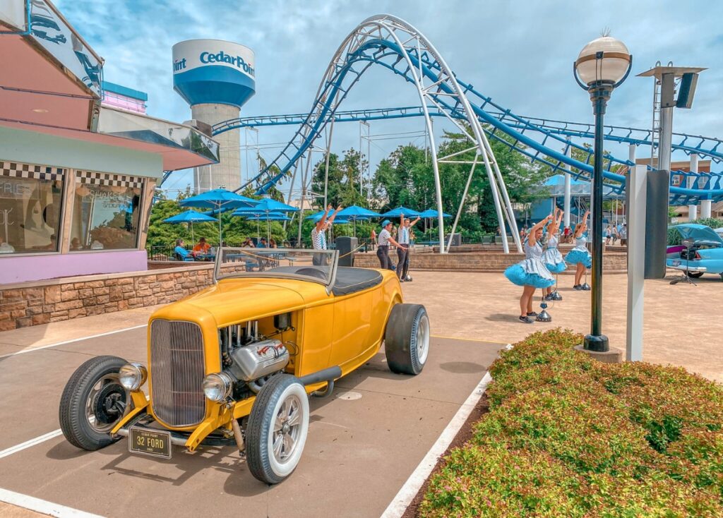 Entertainers perform outside a 50's themed diner at Cedar Point. A blue and white Cedar Point water tower and roller coaster is in the background. In front of the performers is an old yellow Ford. 