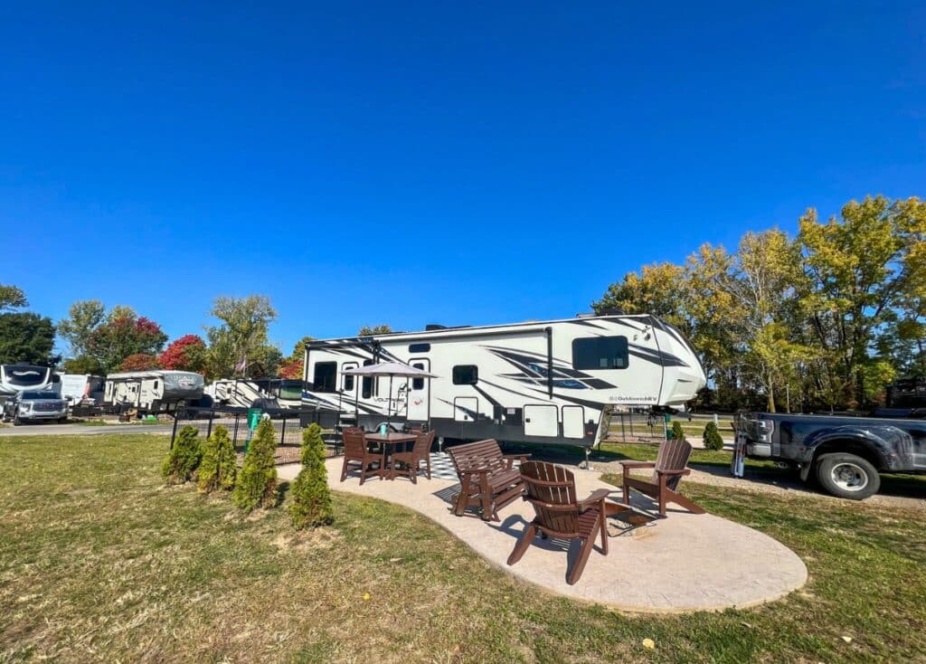 White fifth wheel parked at the Sandusky Cedar Point KOA. The campsite is a pull-through with a patio, brown outdoor furniture, a fire pit, and its own pet pen.