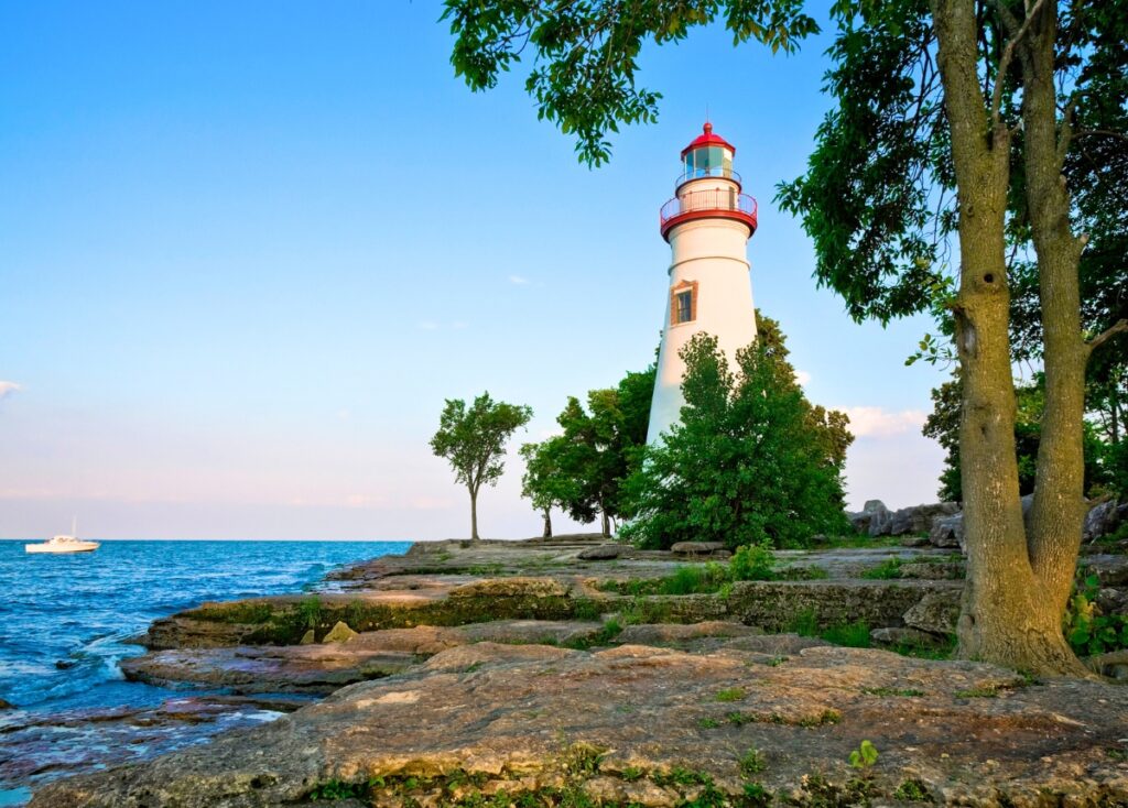 A white and red lighthouse, Marblehead Lighthouse, stands out among trees and rocks overlooking Lake Erie. A boat is out on the water in the distance. 