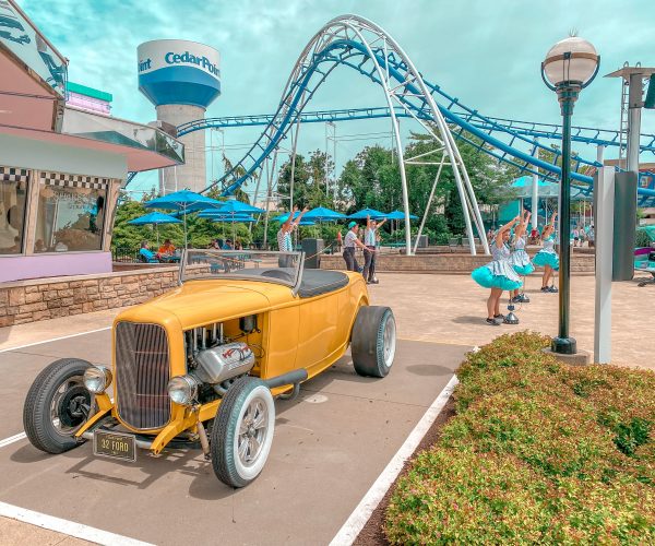Performers at Cedar point outside of diner with old fashioned car. A blue and white water tower with the "Cedar Point" logo is in the background along with a blue and white roller coaster.