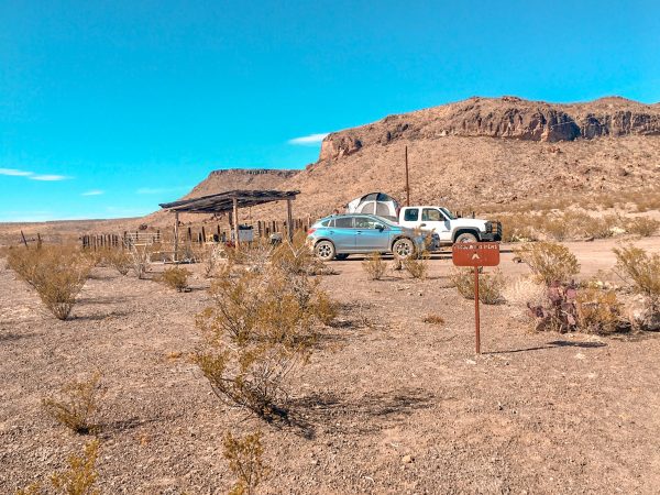 A white truck with a truck tent and a blue Subaru are parked at the Escondido Pens Campsite in Big Bend Ranch State Park. There are small mountains behind the campsite and behind the vehicles is a shaded area with a picnic table. A brown sign marks the name of the campsite.