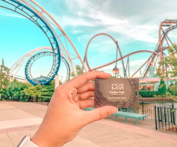 A hand holds up a Cedar Fair Platinum Pass card with the corkscrew roller coaster in the background.