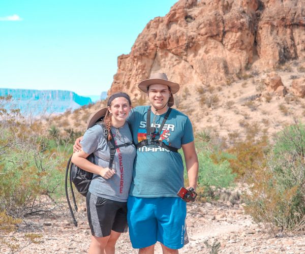 Philip and Megan in front of the Chimneys Big Bend. They're dressed in shorts, t-shirts, hiking backpacks, and Philip is wearing a wide brimmed hat. It's a sunny day and you can see Santa Elena Canyon far off in the distance behind the Chimney.