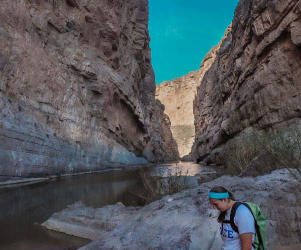 A woman stands with a backpack at the end of the Santa Elena Overlook Trail. The canyon walls tower above while the river runs between them.