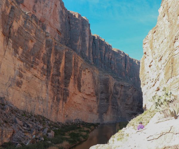 The Rio Grande runs between the towering walls of Santa Elena Canyon. The left side of the canyon is shaded while the right side has sun beating down on it.