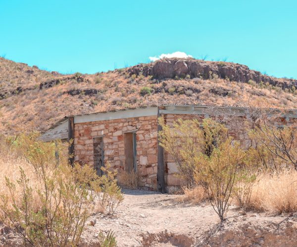 Photo of a building that was once part of the Homer Wilson Ranch in Big Bend. The building is constructed of tan stones, has two doors, and white trim around the top. It's rectangular in shape and has desert shrubbery around it. Behind the home is a small hill with an outcropping of rocks.