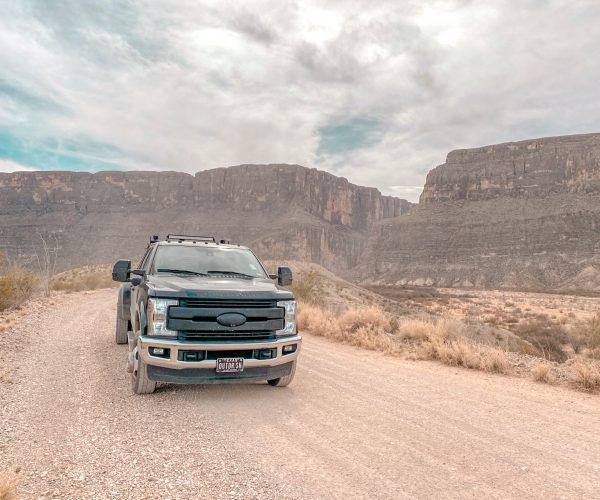 A black Ford F-350 dually drives down Maverick Road, which is a dirt road. Santa Elena Canyon towers in the background behind the truck.