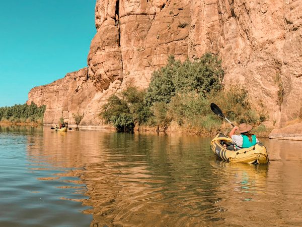 Woman wearing a tan wide brimmed hat and a life jacket paddles a yellow inflatable kayak down the Rio Grande with a canyon wall to her right. Further down the river in front of the woman is another person paddling in a yellow inflatable kayak.