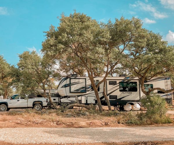 White fifth wheel and truck at campsite parked under trees.
