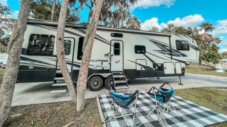 Fifth wheel parked under palm trees with checkered black and white mat setup and camp chairs.