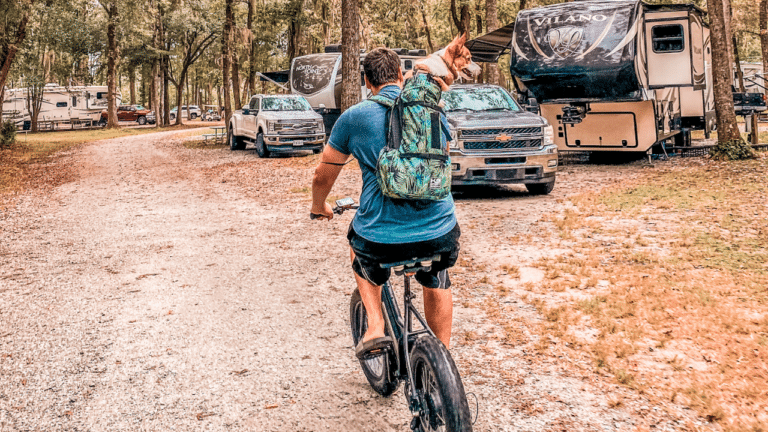 Man riding bike through RV campground while wearing dog backpack with corgi inside.