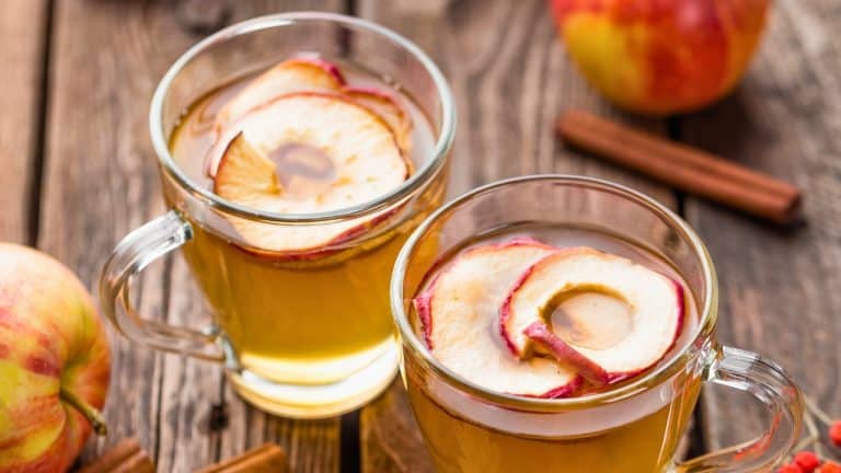 Clear mugs with hot apple cider topped with round apple slices sitting on a picnic table