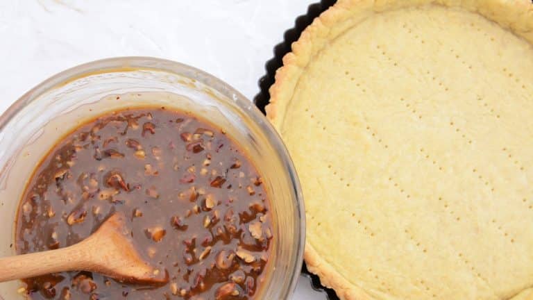 Empty pie crust with bowl of pecan pie filling next to it with wooden spoon resting in bowl