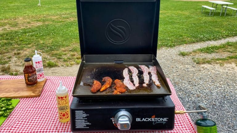 Blackstone griddle sitting atop a picnic table. Bacon and chicken tenders cooking on griddle.
