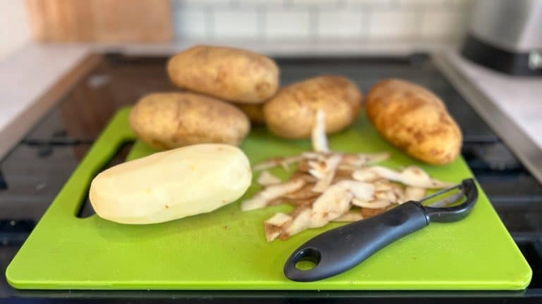 Green cutting board with pile of potatoes sitting on top. One potato is peeled. Potato peels and peeler sit on the cutting board as well