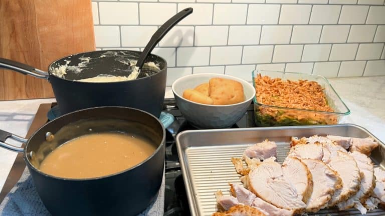 Thanksgiving spread on stove top. Pot of mashed potatoes and pot of gravy, sheet pan holding sliced turkey, green bean casserole, and a bowl of dinner rolls