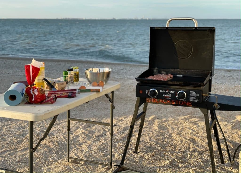 Blackstone griddle on the beach with white folding table setup next to it holding a variety of cooking utensils and ingredients