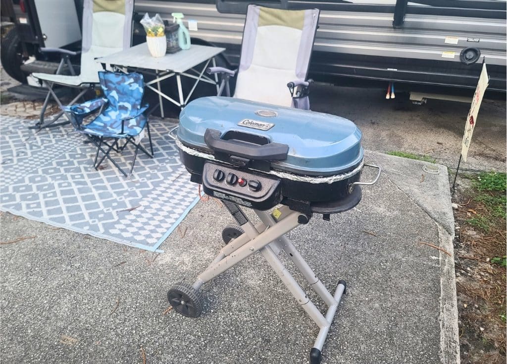 Blue Coleman 225 Portable Propane RV Grill with wheels on patio in front of travel trailer.