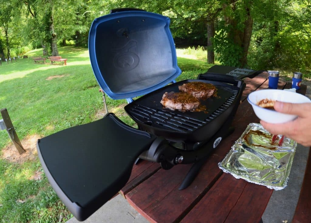 Blue Weber Q1200 RV grill sitting on a picnic table in nature with pork chops being grilled